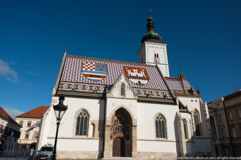 St. Mark’s Church in Zagreb with white walls, Gothic windows, square bell tower, and brightly patterned heraldic tile roof.
