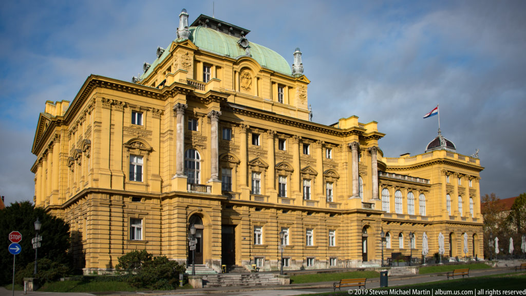Large ochre civic building with rusticated base, tall arched windows, green copper roof, and Croatian flag in Zagreb.