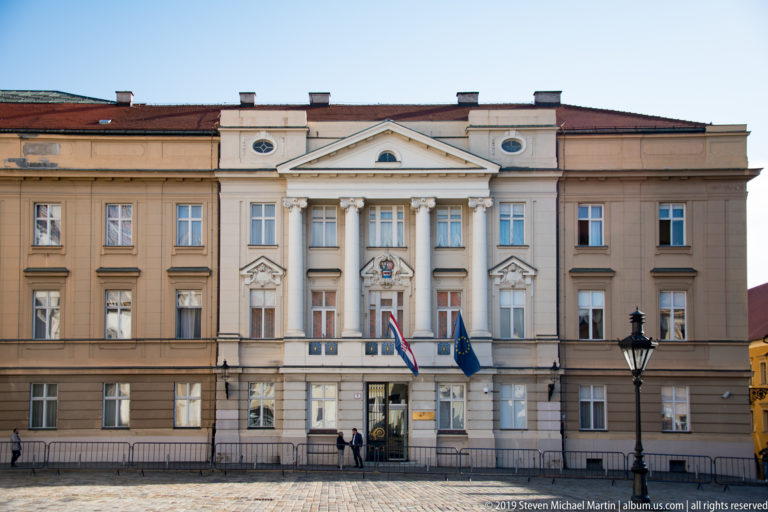 Symmetrical neoclassical government building with columned central entrance, Croatian and EU flags, and rows of tall windows.
