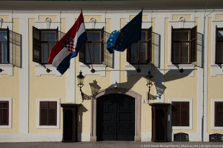 Pale historic façade with dark shutters, arched doorway, wall lanterns, and Croatian and European Union flags.