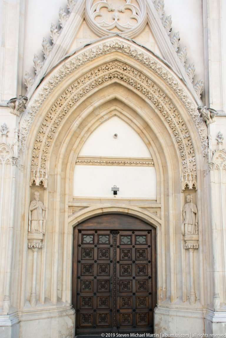 Tall pointed Gothic portal with carved archivolts, side figures, and dark wooden double doors on Zagreb Cathedral.