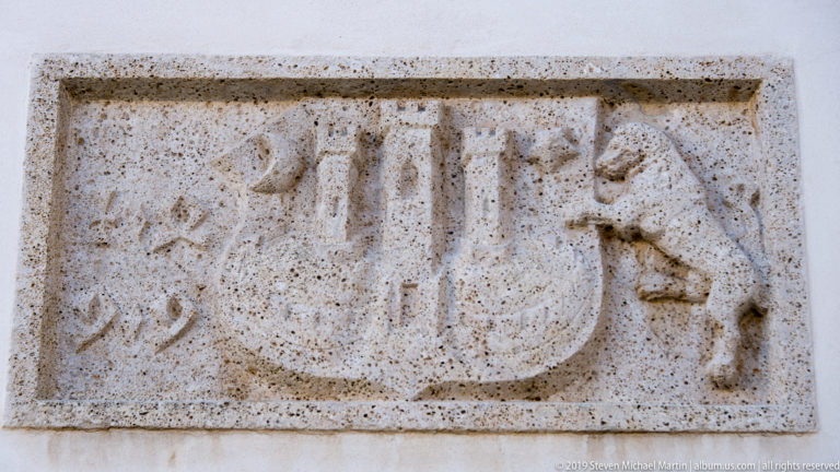 Weathered rectangular stone relief carved with a symmetrical emblem of curved and geometric forms on a pale wall.