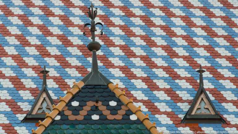 Close view of St. Mark’s Church roof with red, white, blue, and dark glazed tiles behind a small pointed turret.