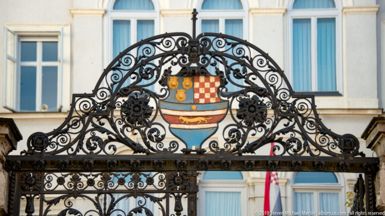 Full wrought-iron gateway with central painted Croatian heraldic shield in front of tall windows on a pale historic building.