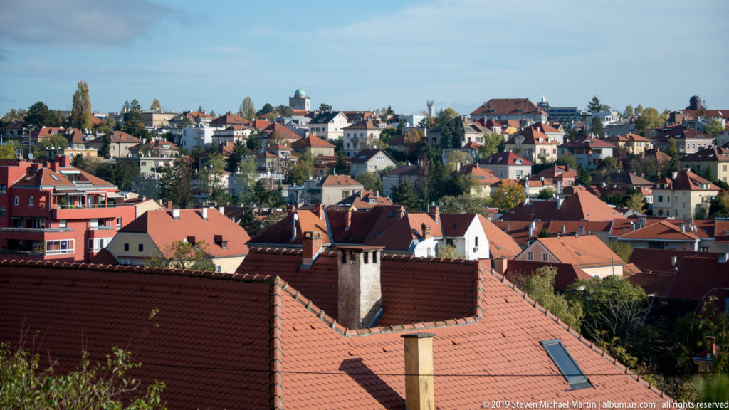Elevated view over Zagreb’s red-tiled Upper Town roofs, chimneys, houses, and distant church towers under a blue sky.