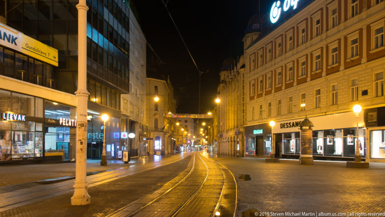 Curving tram tracks run along a quiet Zagreb street lined with illuminated shops and multi-story buildings at night.