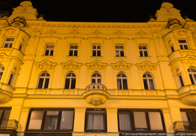 Ornate multi-story historic façade with arched windows, sculpted decoration, curved bays, and central balcony under golden night lighting.