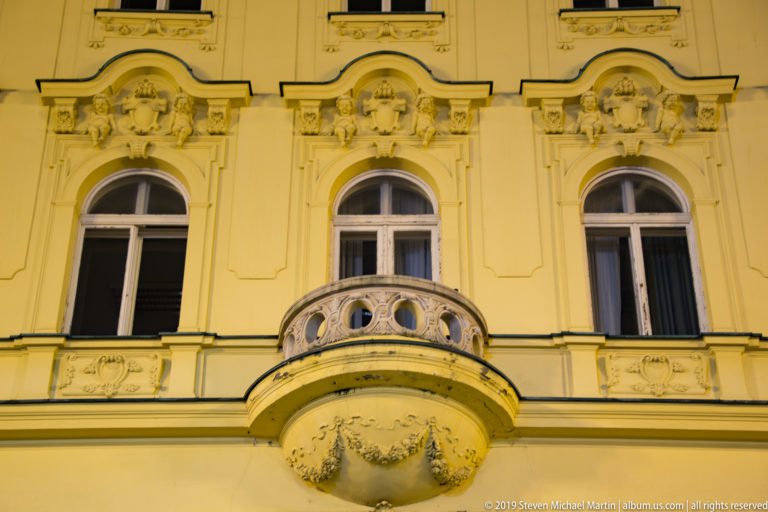 Curved stone balcony beneath tall arched windows on an ornate cream historicist façade illuminated at night.