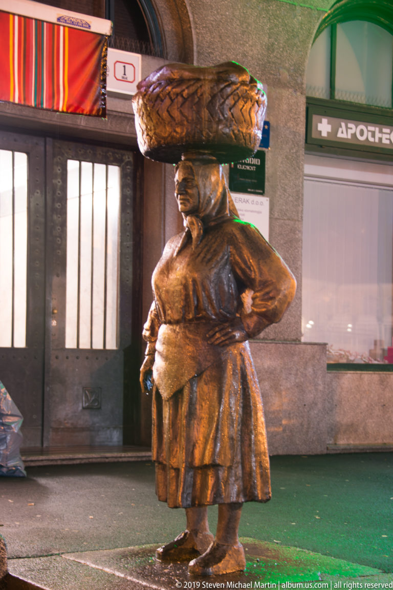 Bronze Kumica Barica market-woman statue carrying a large woven basket on her head outside storefronts at night.