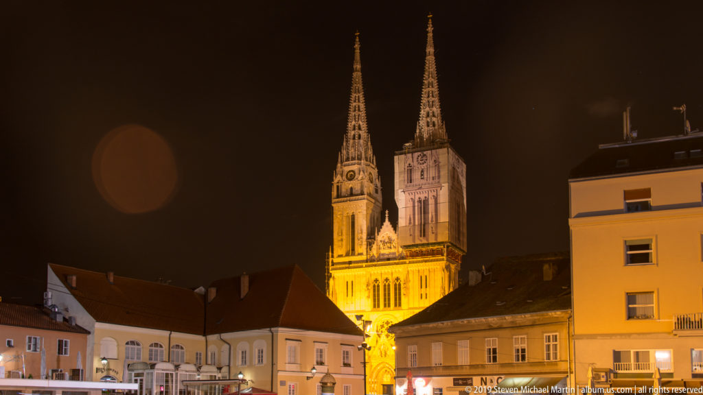 Illuminated twin spires of Zagreb Cathedral rising behind lower historic buildings in a warm-lit nighttime streetscape.