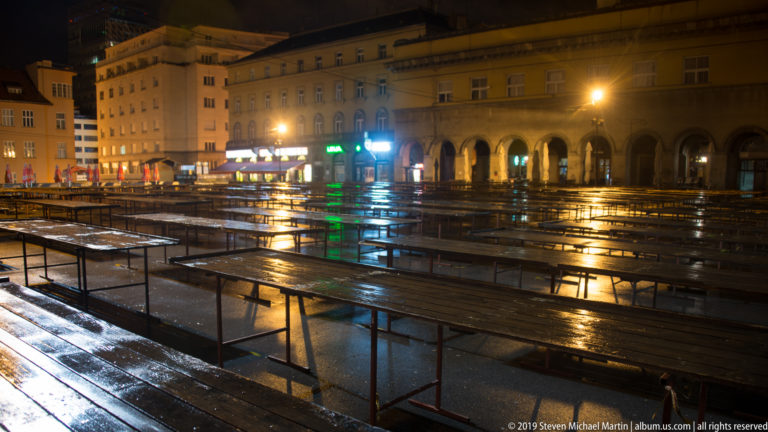Rows of empty wet wooden market tables reflecting lights at night, with arcaded buildings and illuminated storefronts beyond.