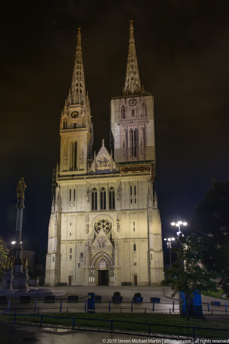 Frontal distant view of illuminated Zagreb Cathedral with twin towers and central portal across an open square at night.