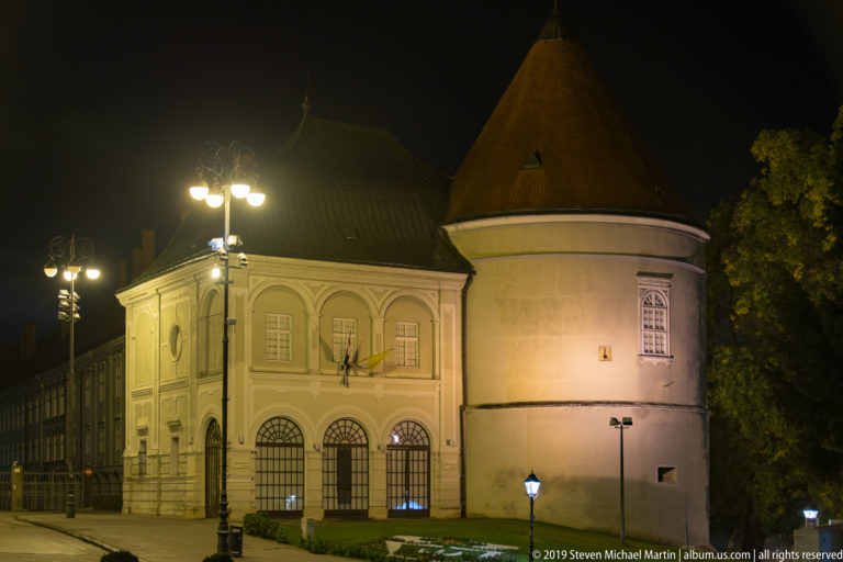 Historic pale building with arched windows adjoining a large round tower with conical roof, illuminated by ornate streetlamps.