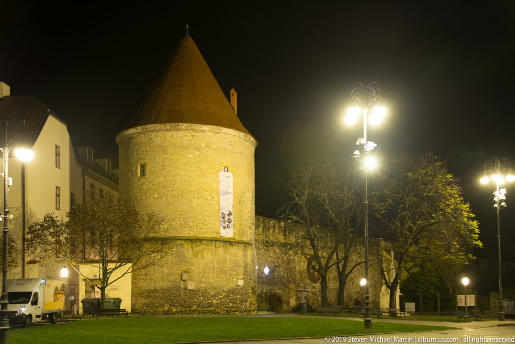 Round defensive tower with conical roof illuminated beside grass, trees, and decorative streetlamps at night.