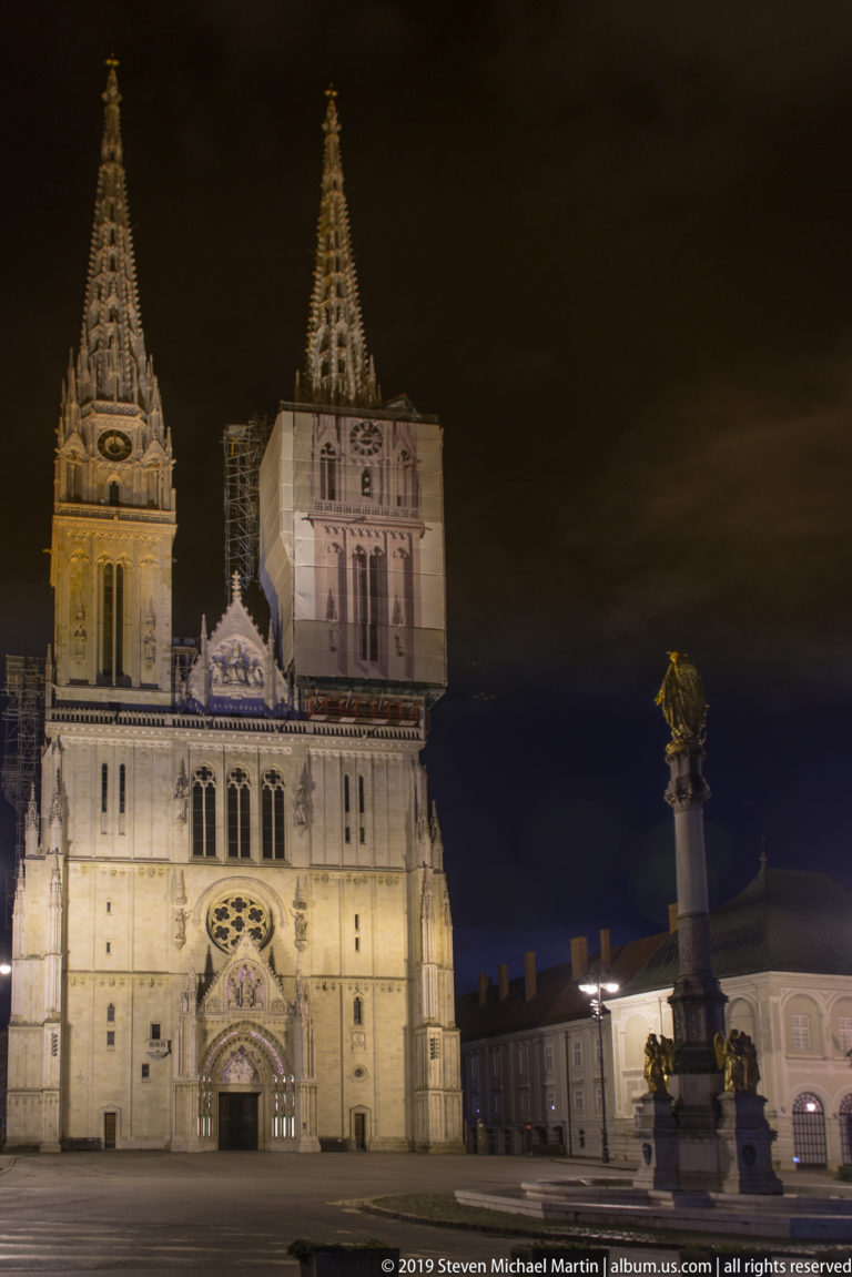 Zagreb Cathedral illuminated at night with its twin towers, while a tall Marian monument stands to the right in the square.
