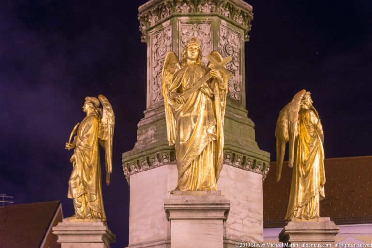 Three gilded winged angel statues arranged around a tall decorated monument base under nighttime lighting.