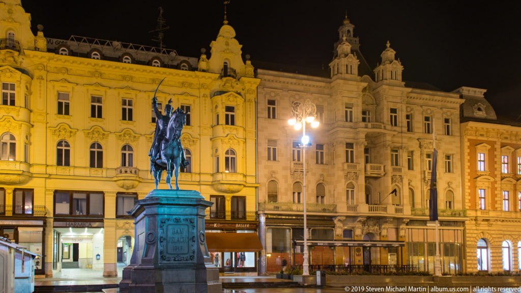 Ban Josip Jelačić monument on its pedestal framed by illuminated historic buildings and shopfronts at night.