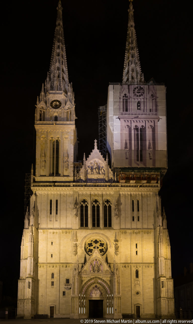 Illuminated west façade of Zagreb Cathedral with twin towers, rose window, ornate portal, and visible work covering on one tower.