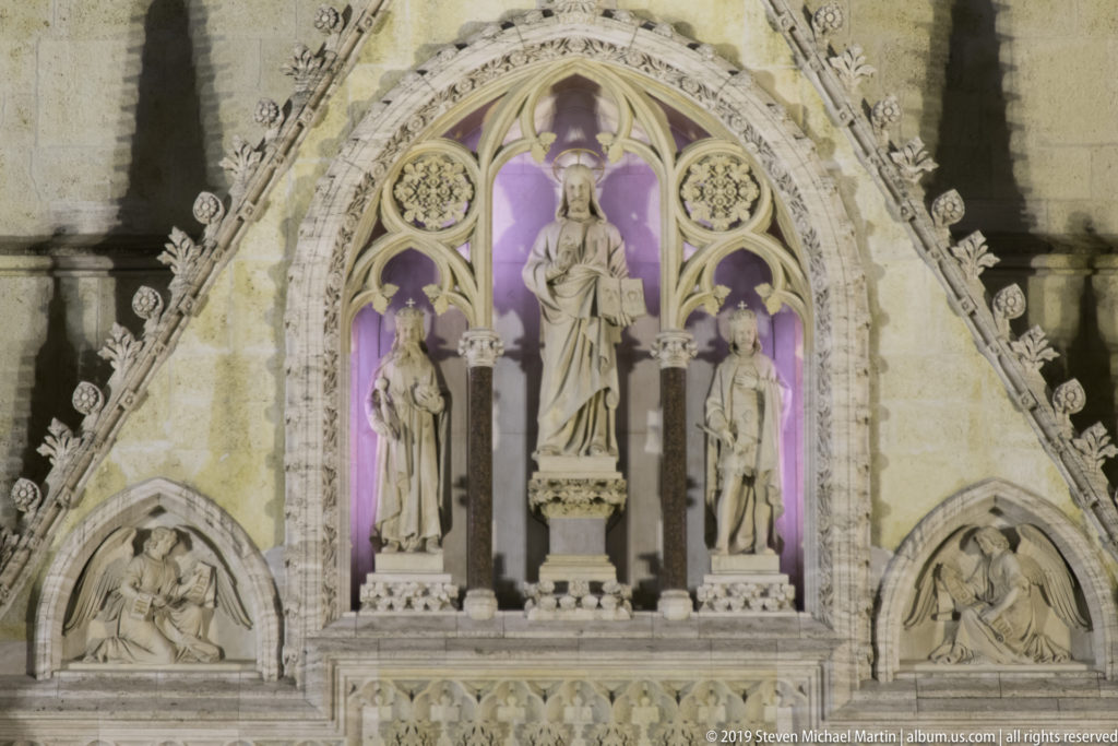 Three saint statues in pointed niches above Zagreb Cathedral’s portal, flanked by relief panels and violet-lit Gothic carving.