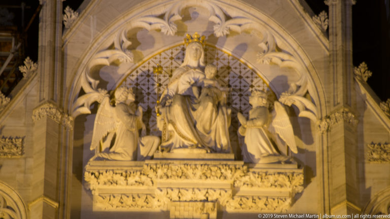Enthroned Virgin and Child flanked by kneeling angels beneath a carved Gothic canopy, illuminated warmly at night.