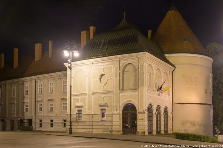 Pale historic building with arched ground floor and steep roof adjoining a round defensive tower on a quiet night street.