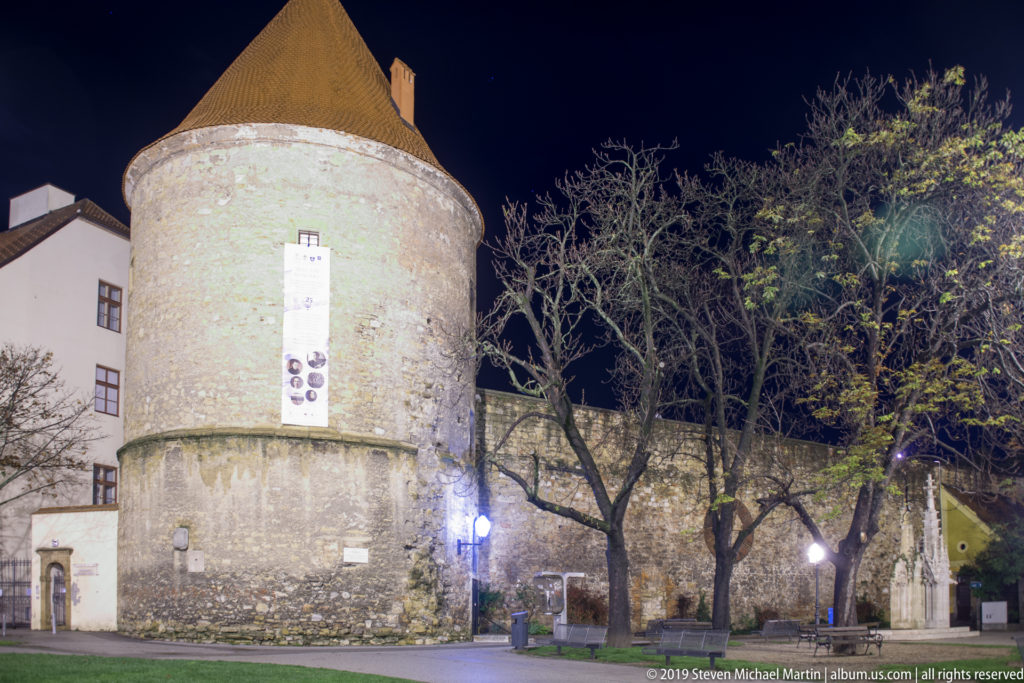 Round masonry defensive tower with rough stone base, pale upper wall, narrow openings, and steep conical roof at night.