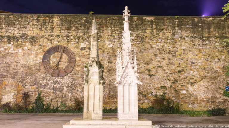 Two slender stone monuments before a historic wall, with a large circular iron clock mounted to the masonry at left.