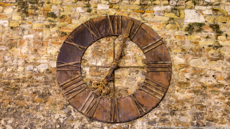 Large rust-toned iron clock with Roman numerals and hands mounted on a rough historic stone wall at night.