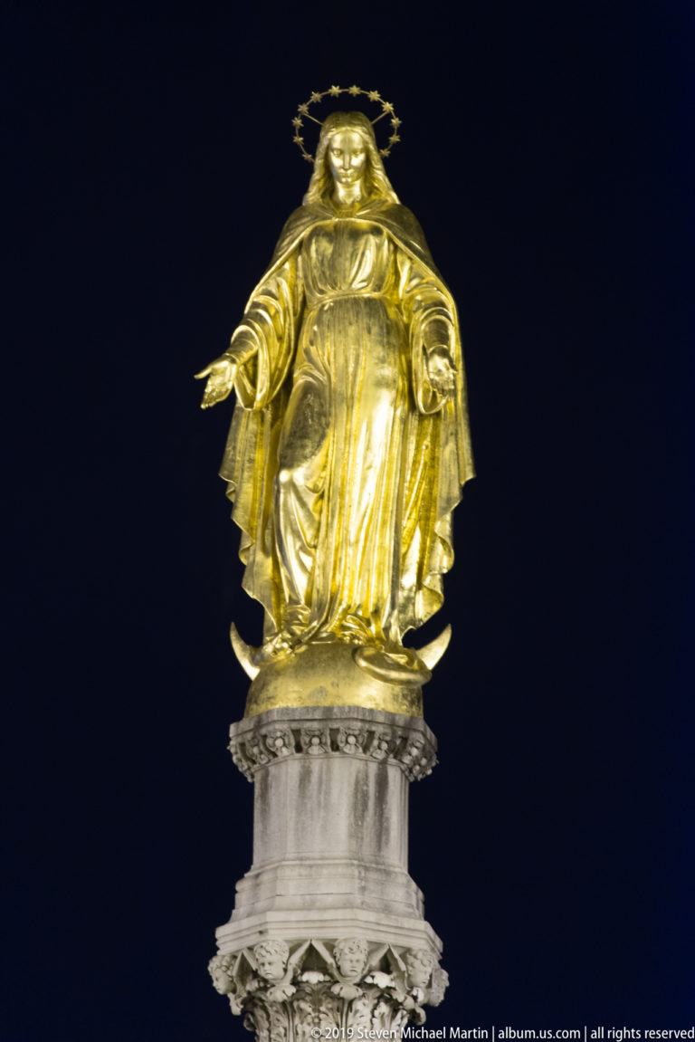 Gilded Virgin Mary statue with star halo and outstretched hands atop a carved column against a black night sky.