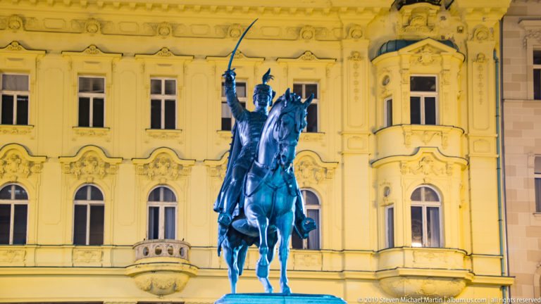 Front view of the illuminated Ban Josip Jelačić equestrian monument with raised sword before an ornate historic building.