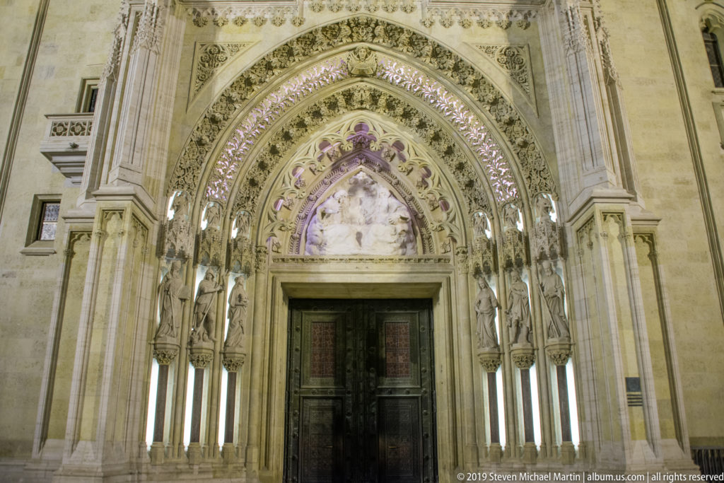 Main entrance of Zagreb Cathedral with dark double doors beneath layered pointed arches, statues, colonnettes, and ornate stone carving.