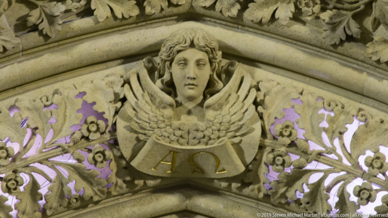 Winged angel carved in stone above Alpha and Omega letters, surrounded by floral Gothic ornament with purple illumination.