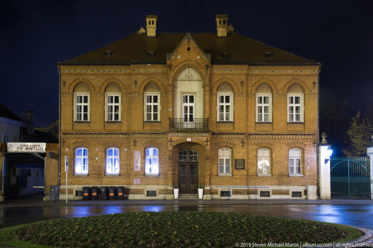 Symmetrical brick civic building with central doorway, tall arched windows, tiled roof, and wet street at night.