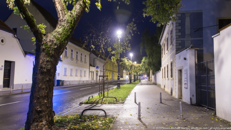 Tree-lined Zagreb street with wet pavement, bicycle racks, bollards, low buildings, and streetlights receding into the night.