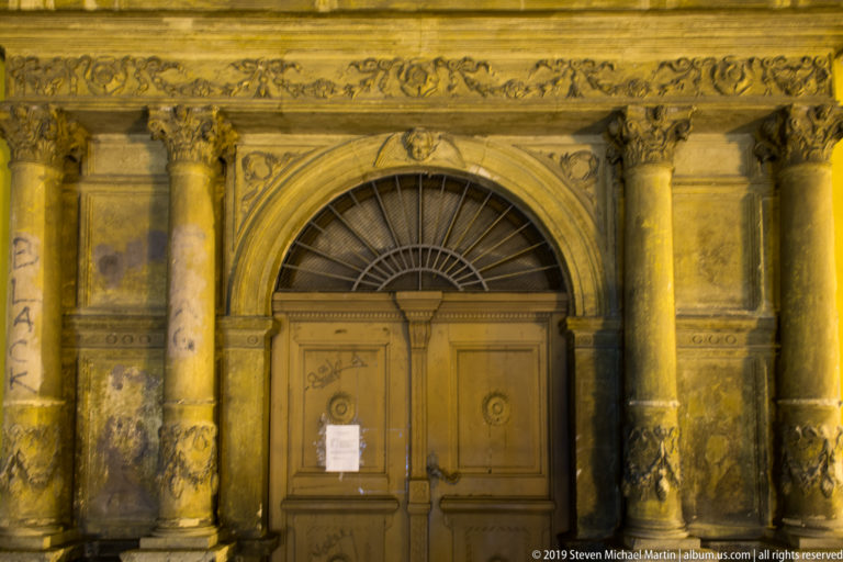Weathered arched stone doorway with paired columns, carved ornament, fanlight, graffiti, and closed wooden doors under warm night lighting.