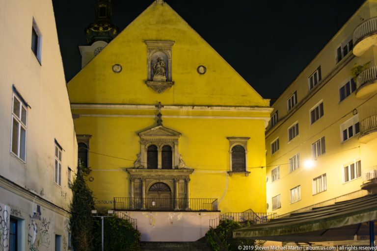 Yellow Baroque church façade at night with stone portal, paired niches, and a devotional image high in the triangular gable.