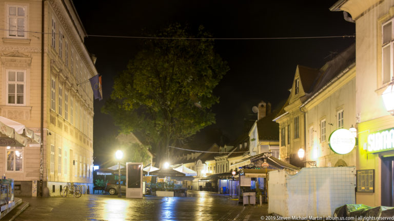 Wide nighttime view along Tkalčićeva Street with a large tree, restaurant canopies, kiosks, historic façades, and wet pavement.