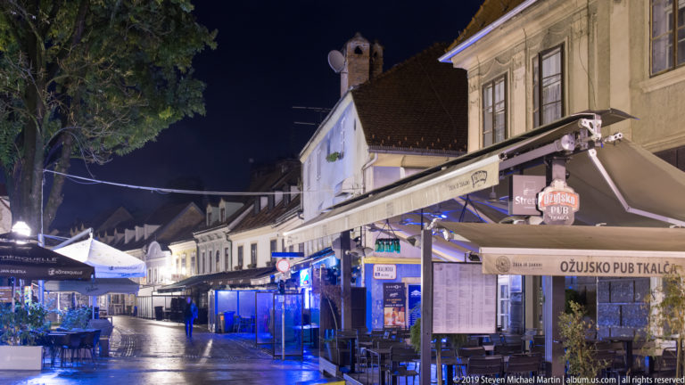 Wet Tkalčićeva Street at night with pub signs, beer awnings, empty café tables, and low historic buildings.