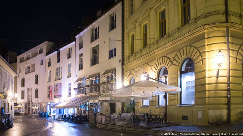 Curving Zagreb street at night with restaurant umbrellas, outdoor seating, a rounded historic corner building, and modern façades.