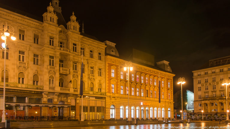 Illuminated multi-story historic buildings and street lamps lining Ban Jelačić Square beneath a dark night sky.