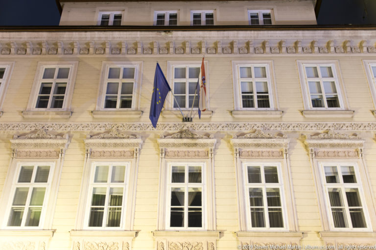 Ornate pale civic façade at night with Croatian, European Union, and central flags above rows of tall windows.
