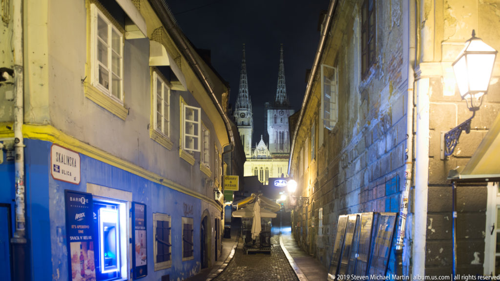 Narrow Zagreb lane at night leading toward the illuminated twin spires of Zagreb Cathedral in the distance.