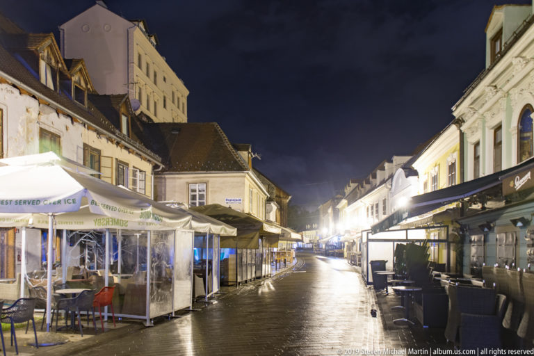 Empty café terraces and awnings line wet Tkalčićeva Street beneath bright nighttime lights in Zagreb.