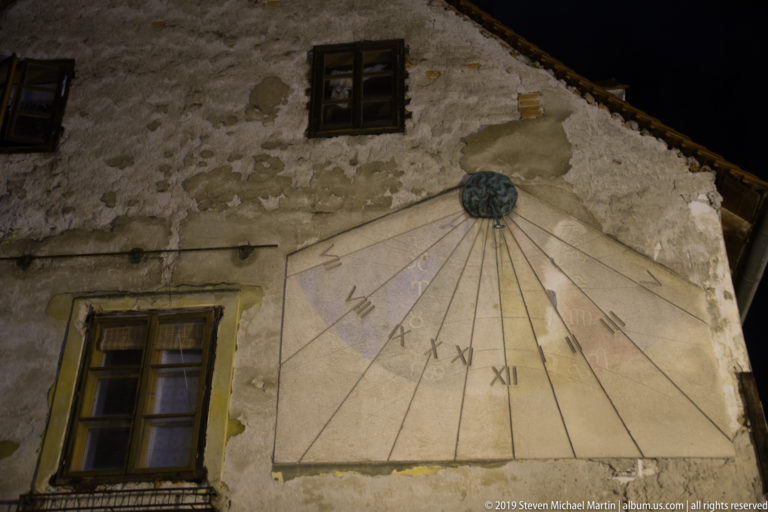 Large painted wall sundial with Roman numerals and radiating lines on a weathered plaster façade at night.