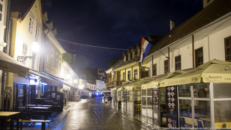 Wet pedestrian lane lined with empty restaurant terraces, awnings, umbrellas, and historic façades on Tkalčićeva Street at night.