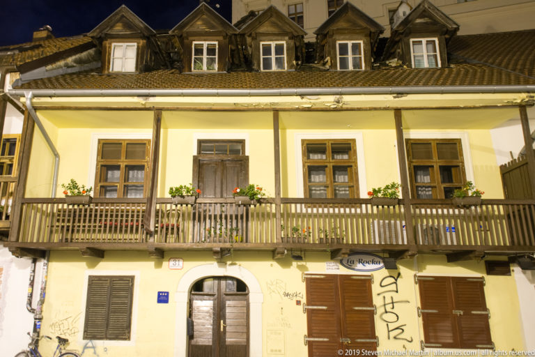 Long historic building with wooden balcony, flower boxes, dormer windows, closed shops, and graffiti at night.