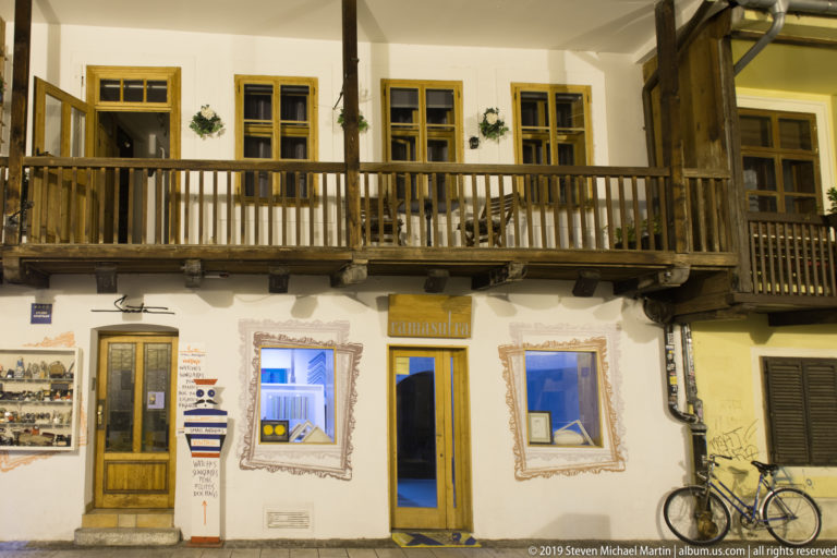 Traditional two-story building with long wooden balcony, yellow-trimmed windows, shopfronts, and a bicycle on Tkalčićeva Street at night.