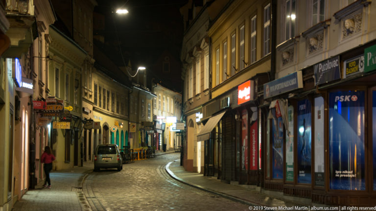 Narrow curving Zagreb street at night with shops, signs, a parked van, and a lone pedestrian beneath streetlights.