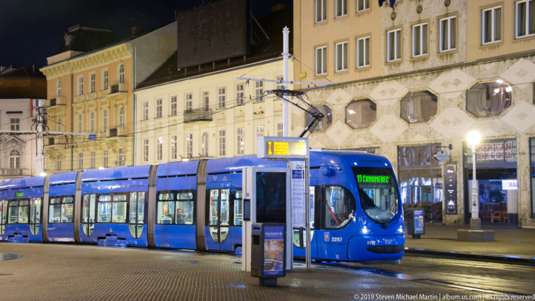 Blue articulated ZET tram stopped beneath overhead wires on an illuminated central Zagreb street at night.