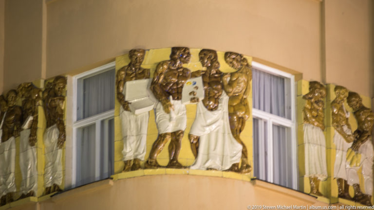 Polychrome high-relief human figures wrap around a projecting corner window on a Zagreb building.